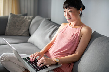 Smiling Asian woman working on laptop from home sofa