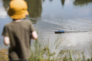 A 5-year-old boy in a yellow panama hat launches a radio-controlled boat in a pond. Children's fun...