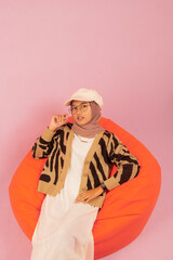 Portrait of beautiful young woman in hijab wearing glasses and hat posing for camera sitting on orange bean bag with angle from above, pink isolated studio background