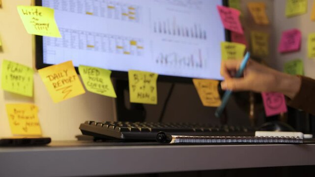 Close-up of hand of woman placing colorful papers on her computer screen, working late at night, walls covered in sticky notes. Selective focus. Time organization and planning