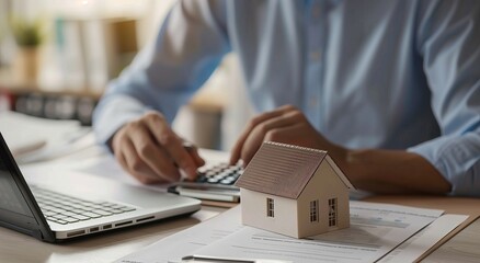 Fototapeta premium Real estate agent sits at his desk, looking over the realistic house model on top of papers with a calculator in front and holding an envelope in one hand while taking notes