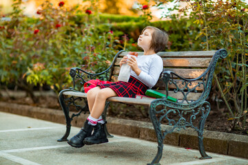 A little girl, a schoolgirl, is sitting on a bench and eating a sandwich