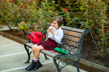 A little girl, a schoolgirl, is sitting on a bench and eating a sandwich