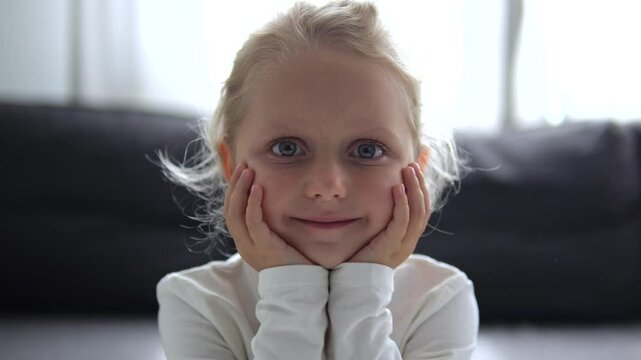 Charming blond girl propped up her chubby cheeks with her hands and gives radiant smile. Happiness shines in eyes, carefree smile spreads across face. Charming child smile radiates joy and spontaneity