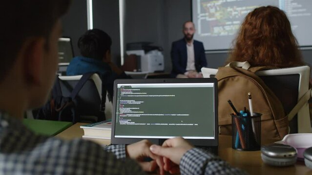 Medium rear shot of group of teenage school students sitting at desks with laptops with programming code, listening to Middle Eastern male teacher explaining new subject next to projector screen