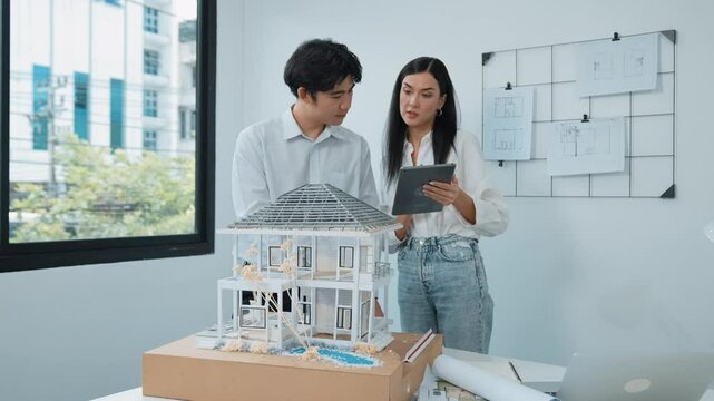 Beautiful young caucasian project manager brief asian engineer by using tablet while engineer measuring house model on meeting table with blueprint, architectural equipment scatter around. Immaculate