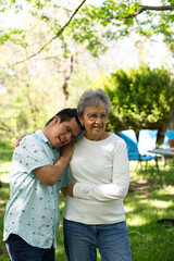 A young man and an older woman embrace in a park. The man is wearing a blue shirt and the woman is wearing a white shirt. The scene is peaceful and comforting.