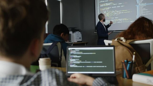 Medium full rear shot of group of children sitting at desks with laptops in classroom, listening to Middle Eastern male teacher pointing to computer code on projector screen and explaining new subject