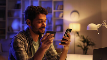 Smiling pretty young Arabian brunette boy holding mobile phone and credit card sitting at the table. Happy man makes online shopping using mobile payment. E-banking app service.