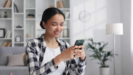 A young African American girl sits at the table in the living room, smiles, chats with friends on a mobile phone, happily taps the screen, reads happy news.
