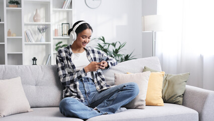 Happy young girl with white headphones chilling and listening her favorite song. African American brunette woman sitting on sofa relaxing after work, using smartphone to play music tapping on screen.