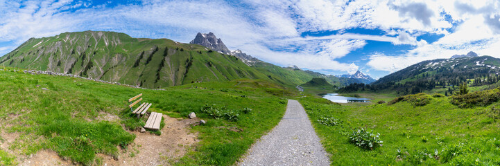 Panorama with a park bench on a hiking trail with a view of a lake and the surrounding mountains in...