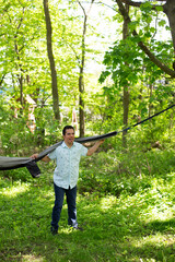 A young man holds a large black and gray hammock. The hammock is suspended between two trees. The man smiles and enjoys the fresh air.