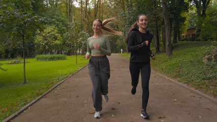 Caucasian young women girls runners joggers sport ladies friends jogging together in park smiling two fit females running enjoying healthy lifestyle morning jog run workout morning activity outside