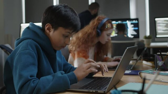 Medium shot of focused young teenage Indian boy sitting at desk in classroom at programming lesson, typing computer code on laptop, and teacher checking on project of red-haired girl in background