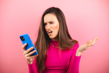 Mad and shock young brunette beautiful woman using mobile phone isolated on pink background. Yelling unhappy woman hold mobile phone. Disappointed sad upset lady horrified impressed news. WTF. Oh no.