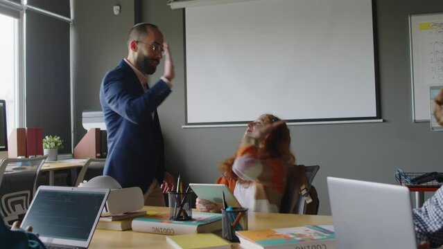 Medium shot of young male computer science teacher checking finished project of female student developing mobile application on tablet in programming class, commending on it and giving girl high five