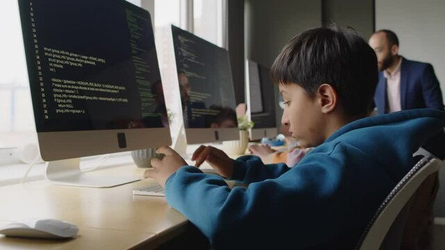 Medium side shot of young biracial Indian boy sitting at desktop computer at programming lesson at school, writing code on black screen, and Middle Eastern teacher walking around class and checking