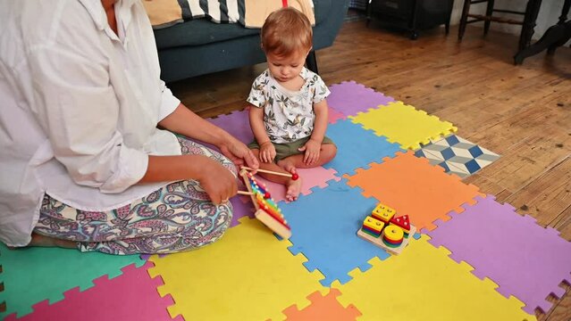 Happy mother and baby enjoying time together with colorful educational toys on a multicolored puzzle carpet. The baby focuses on playing with a xylophone while mother supports and smiles lovingly