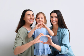 Young lesbian couple with adopted little girl making heart gesture on light background