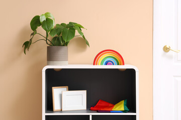 Shelf unit with LGBT flag, toy rainbow and plant near beige wall in room