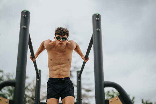 Shirtless young athlete wearing sunglasses, performing a dip exercise on parallel bars in an outdoor gym environment.