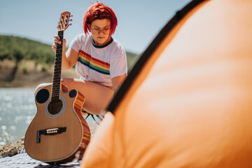 Lesbian girl with red hair sitting by a lake, holding a guitar, and enjoying outdoor relaxation near a tent.