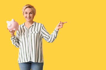 Beautiful mature woman with piggy bank pointing at something on yellow background