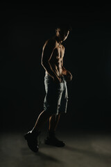 Young shirtless man in denim shorts and boots posing in dark atmospheric studio lighting.
