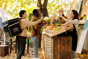 Female courier waving goodbye to small farm business owner, ready to deliver organic produce to customers. Local vendor greeting young lady carrying thermal bag for delivery of bio goods.