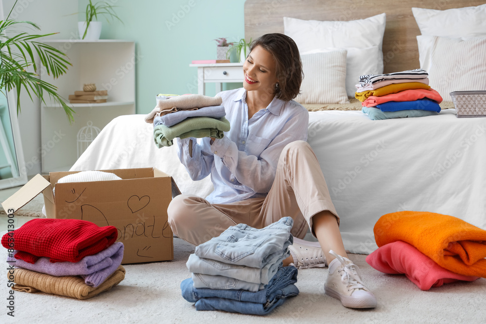 Wall mural young pretty woman packing different clothes in donation cardboard box on floor at home