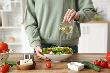Young woman pouring oil into bowl with fresh vegetable salad in kitchen