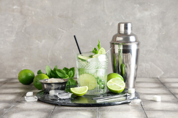 Glass of fresh Mojito cocktail with limes and shaker on grey tile table