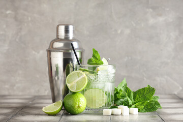 Glass of fresh Mojito cocktail with limes and shaker on grey tile table
