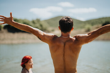 Man with arms wide open at a scenic lake on a sunny day, enjoying the outdoors with a woman in the background.
