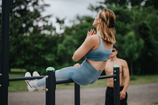 Woman practicing calisthenics workout on parallel bars in an outdoor park with a trainer's guidance, focusing on fitness and strength training.