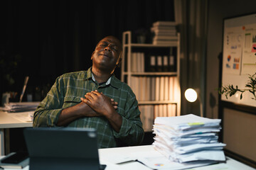 African American Man in Office Holding Chest in Emotional Moment with Stack of Papers and Laptop