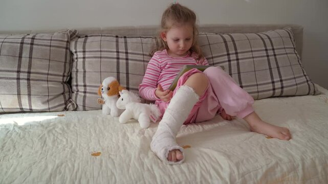 Little girl with broken foot, having plaster cast sitting in her room calmly and reading a book 