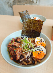 A delicious bowl of ramen featuring tender grilled chicken, mushrooms, perfectly cooked soft-boiled eggs topped with sesame seeds, and garnished with fresh green onions and crispy nori
