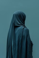 A person wearing a blue scarf is standing in front of a blue wall. The scarf is long and flowing, and the person appears to be looking off into the distance