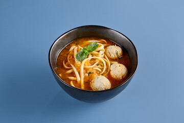 Delicious Chicken Soup with Meatballs and Noodles in a Black Bowl on Blue Background