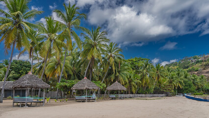 Sun loungers under thatched canopies stand in a row on the sandy beach. The boat is at the shore. Lush palm trees against a background of blue sky and clouds. Madagascar. Nosy Be 