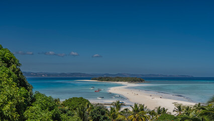 Beautiful postcard view. A sandbar meanders towards a tropical island with green vegetation. Tiny silhouettes of people on the beach. Boats in the aquamarine ocean. Palm leaves in the foreground.