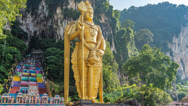 Many people climb the steps of the rainbow-colored staircase to the temple. Lush vegetation on the slopes of a sheer cliff. In the foreground- a giant golden statue of the god Lord Murugan. Batu caves