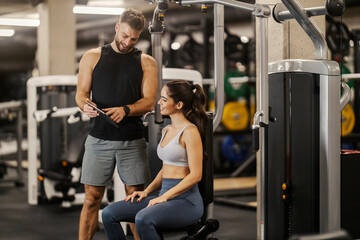 Fit sportswoman sitting at machine in a gym and having consultations with her fitness trainer.