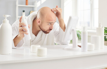 Concerned balding young man in white bathrobe sitting at table with sprays and shampoos for damaged roots and premature hair loss issue treatment, looking in mirror, and rubbing his top and forehead