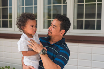 An Asian man in his 50s proudly bonding with his youngest son outdoors. The father is carrying his child near a building with a windowed facade.