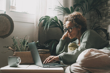 Blonde woman with curly hair using laptop sitting on sofa at home. 45 year old serious middle aged...
