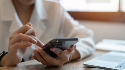Fototapeta premium A woman is using her cell phone while sitting at a desk with a laptop. She is looking at the screen intently, possibly checking her email or browsing the internet. Concept of productivity and focus