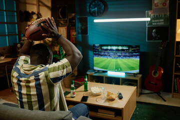 Unrecognizable young African American football fan holding ball above his head watching match on TV in living room, copy space
